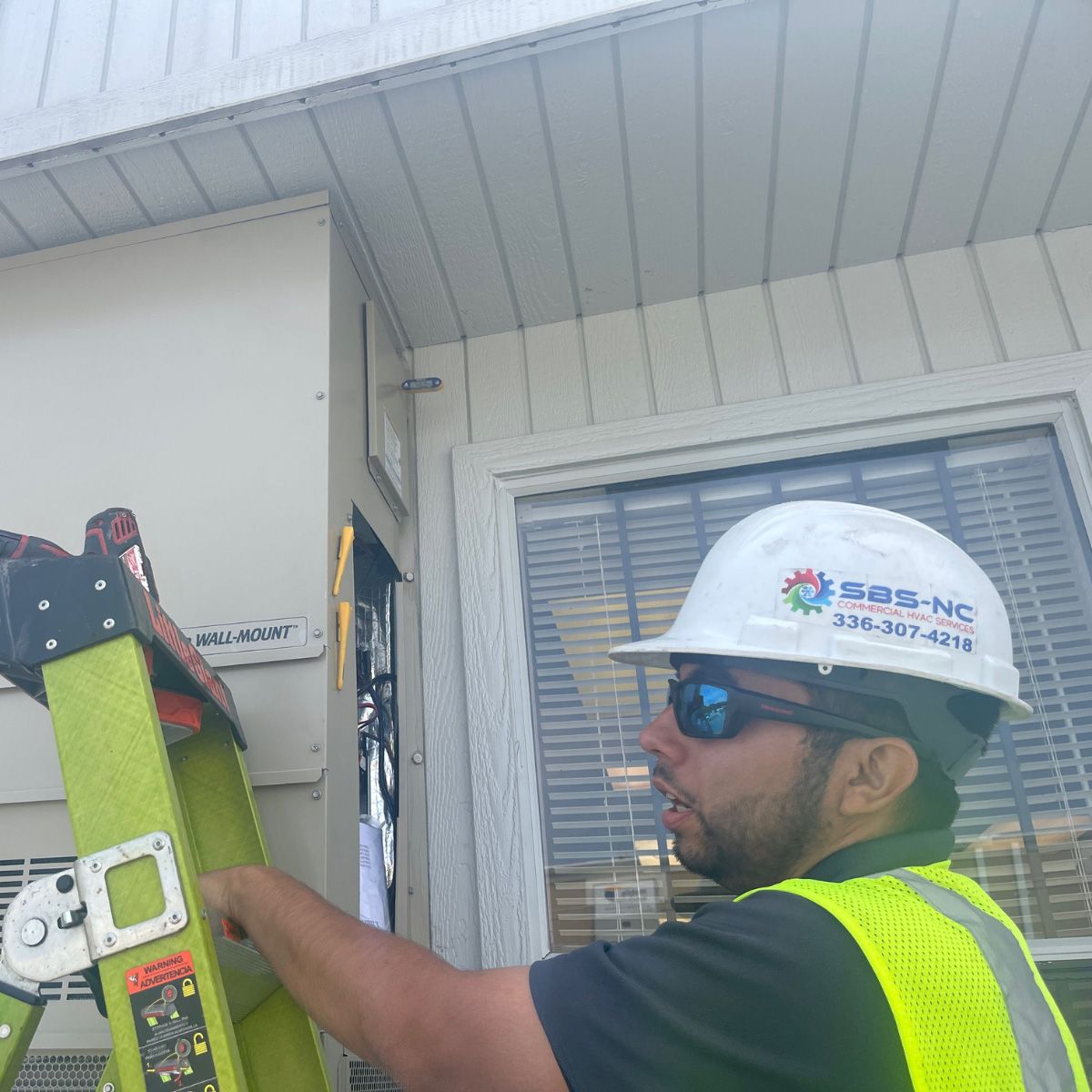 Worker in hard hat and safety vest adjusting equipment beside a building window.