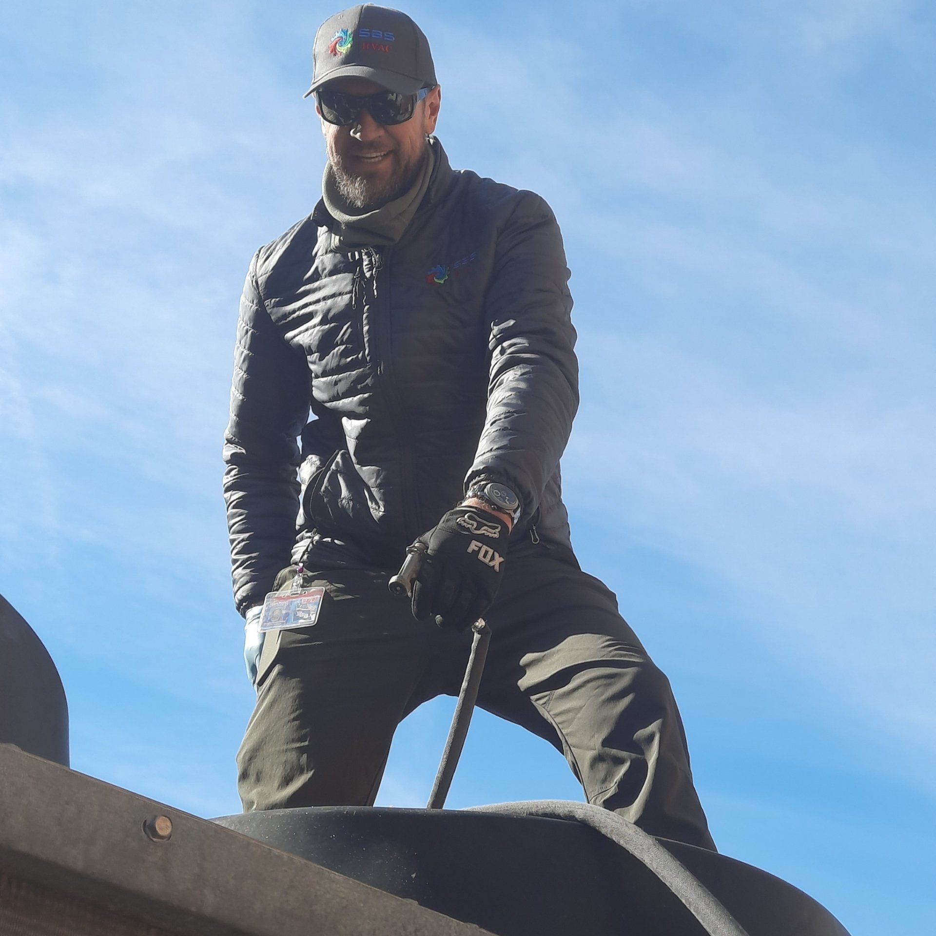 Person in dark jacket and cap standing on a rooftop against a bright blue sky.
