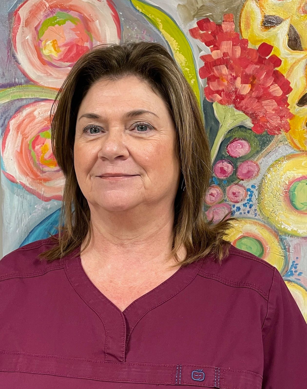 A woman in a purple scrub top is standing in front of a painting of flowers.