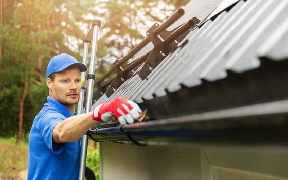A worker in a blue uniform and gloves cleans debris from a house gutter while standing on a ladder.