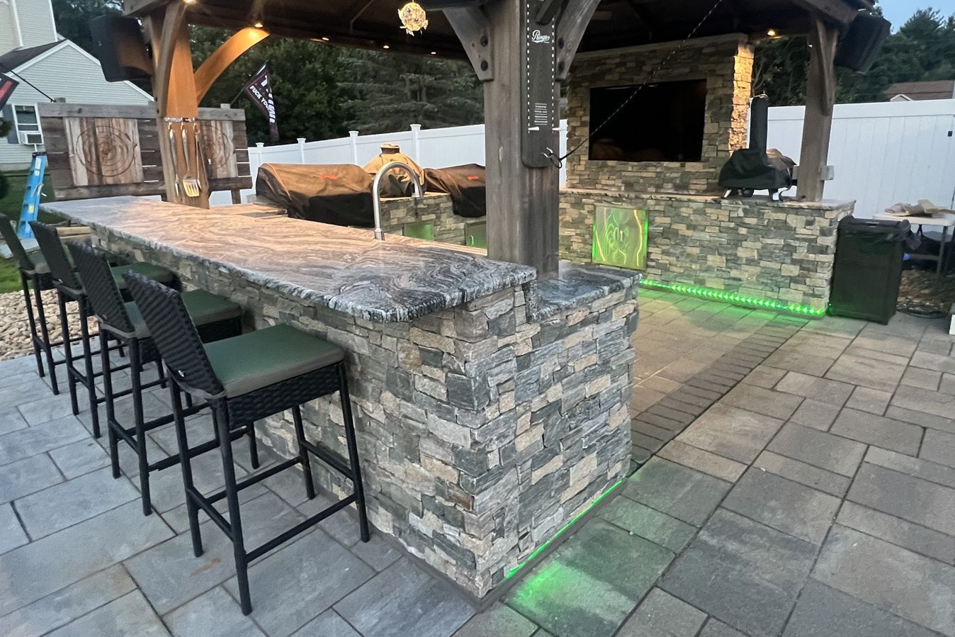 Outdoor stone patio bar with stools under a wooden pergola, featuring an integrated grill, TV, and green floor lighting.