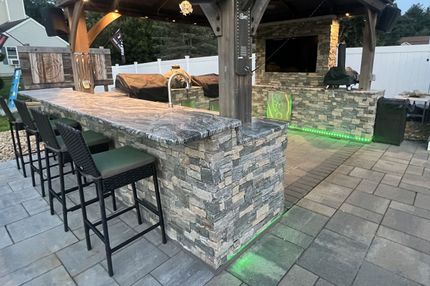 An outdoor kitchen with stone counters, bar seating, a TV, and green LED accent lighting under a wooden gazebo.