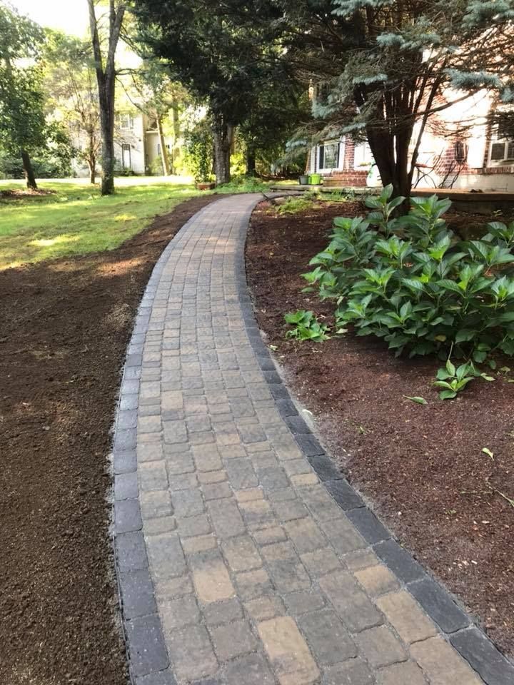 A curved stone paver walkway leads through a landscaped yard with dark mulch and green shrubs.