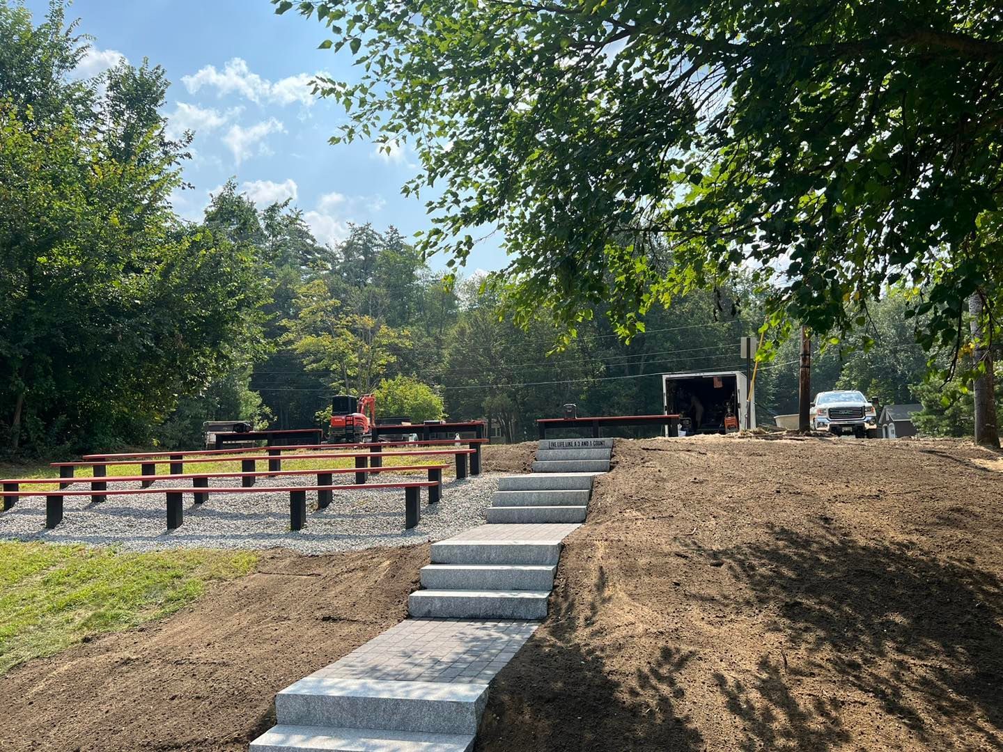 A set of grey stone steps leading up a dirt hill to a gravel seating area with wooden benches among trees.