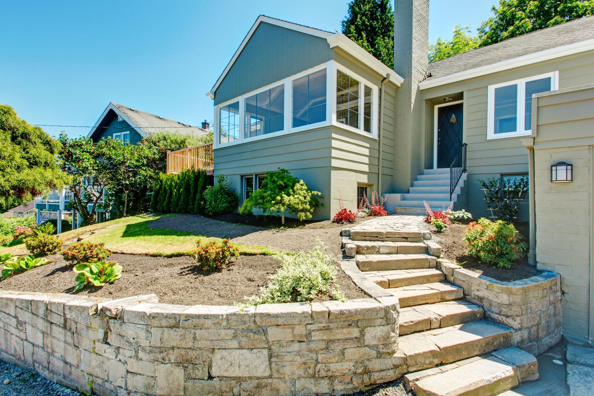 A light green house with a stone retaining wall and stone stairs leading to the front entrance on a sunny day.