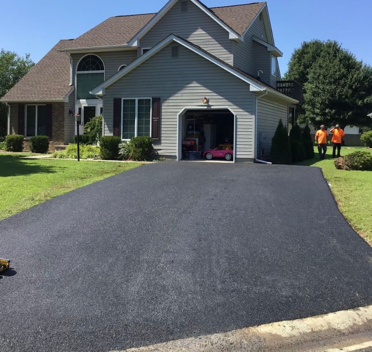 Freshly paved black driveway leading to a gray suburban house with workers in orange vests nearby