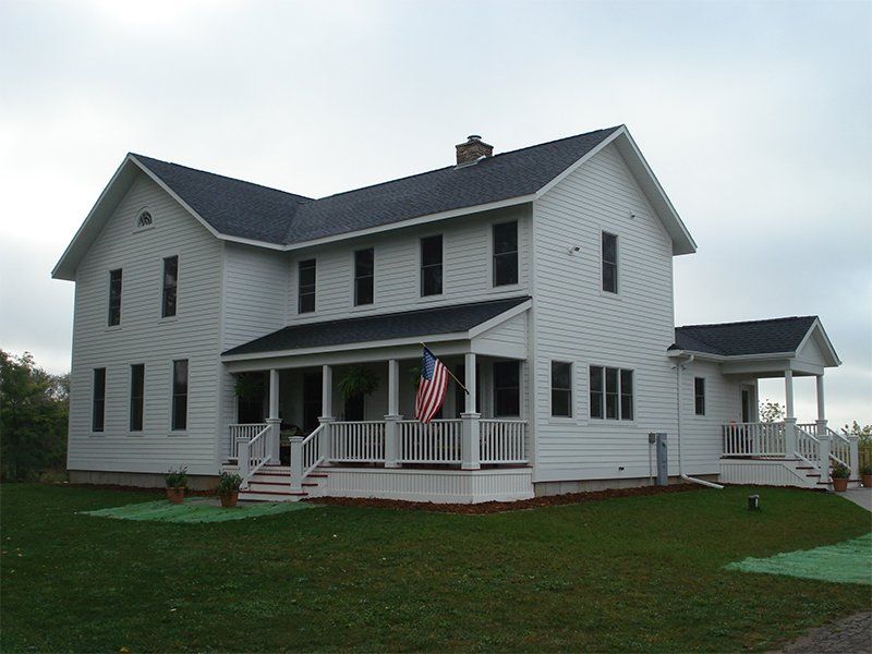White two-story farmhouse with a porch, American flag, and large lawn under an overcast sky