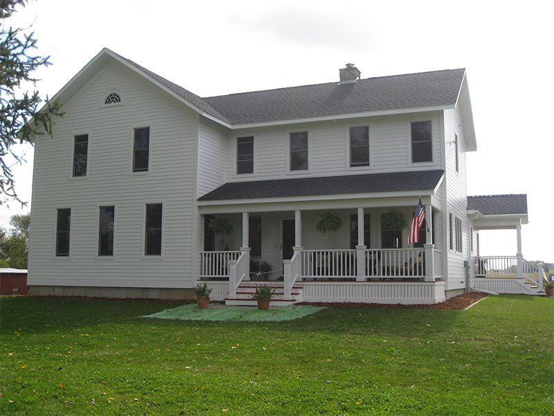 White two-story farmhouse with front porch, green lawn, and an American flag near the entrance
