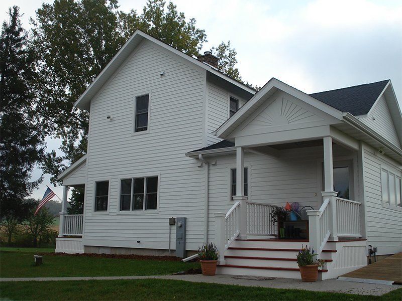 White two-story house with a front porch and steps, set on a grassy lot with trees nearby