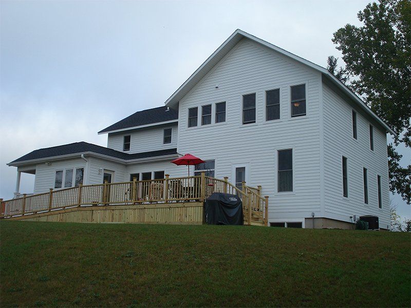 White two-story house with a wooden deck and red umbrella on a grassy hill under a cloudy sky