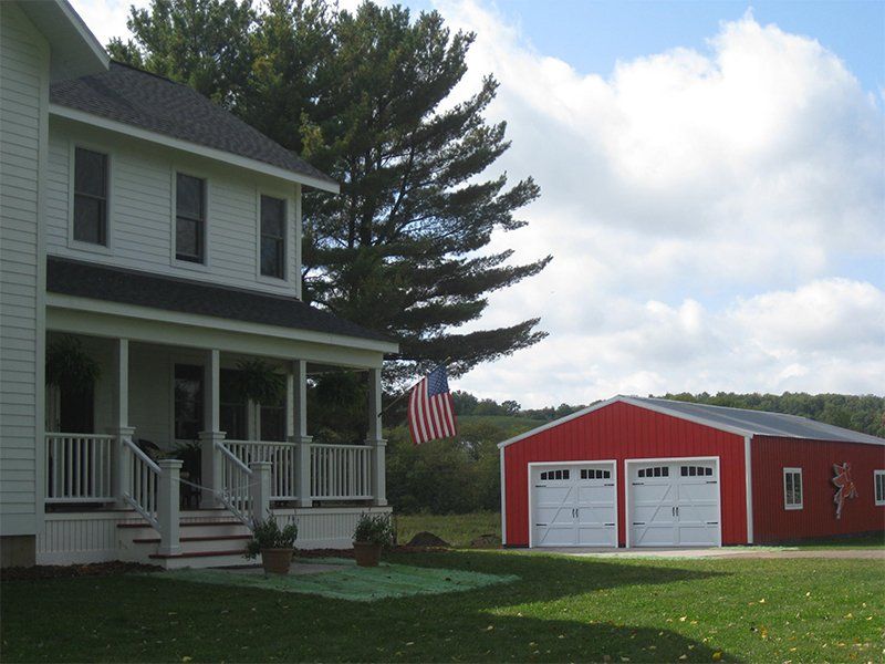 White farmhouse with porch beside a red garage and American flag on a grassy lawn under cloudy skies