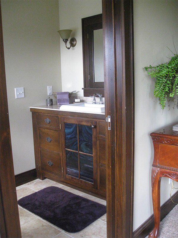 Bathroom vanity with dark wood cabinet, white sink, mirror, wall sconce, and dark purple bath mat.