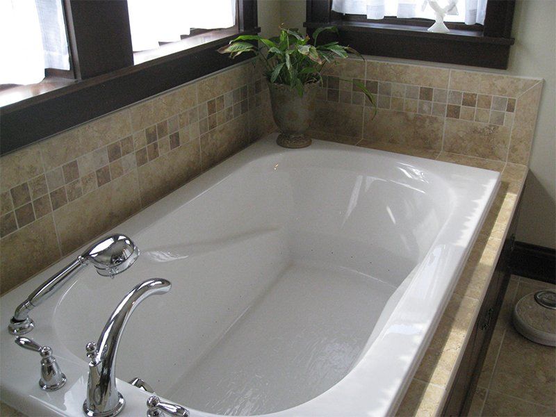 White bathtub with chrome faucet in a tiled bathroom corner, beside windows and a potted plant