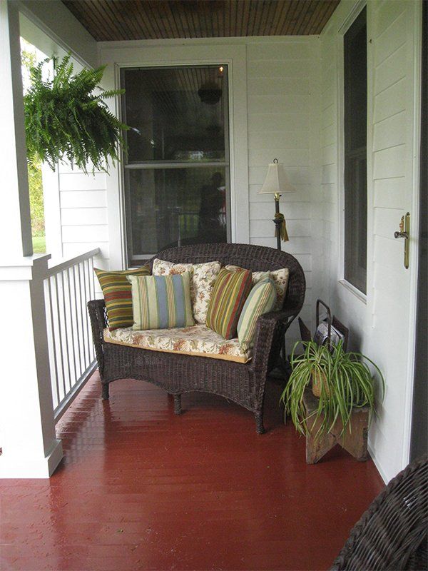 Covered porch with red floor, wicker chair with striped cushions, hanging fern, and potted plant by a white door