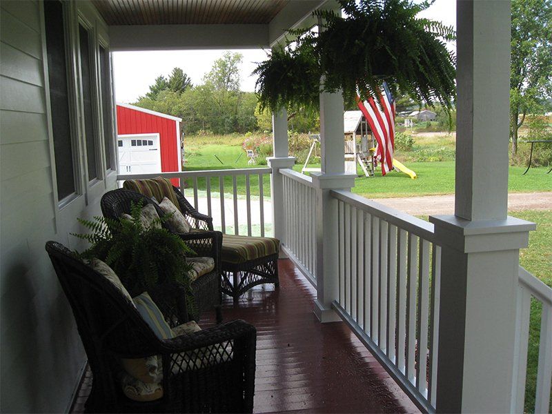 Covered front porch with white railing, wicker chairs, potted ferns, and an American flag overlooking a green yard.