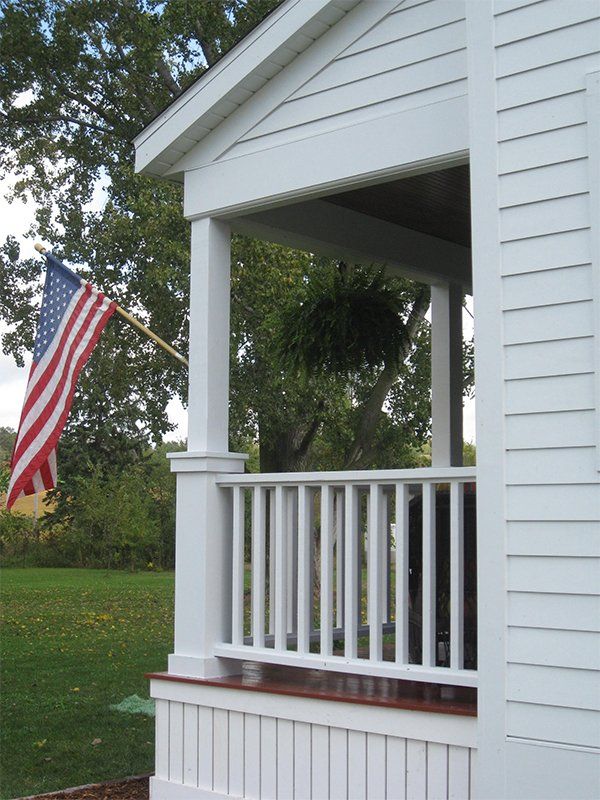 White house porch with railing and an American flag flying beside a grassy yard.
