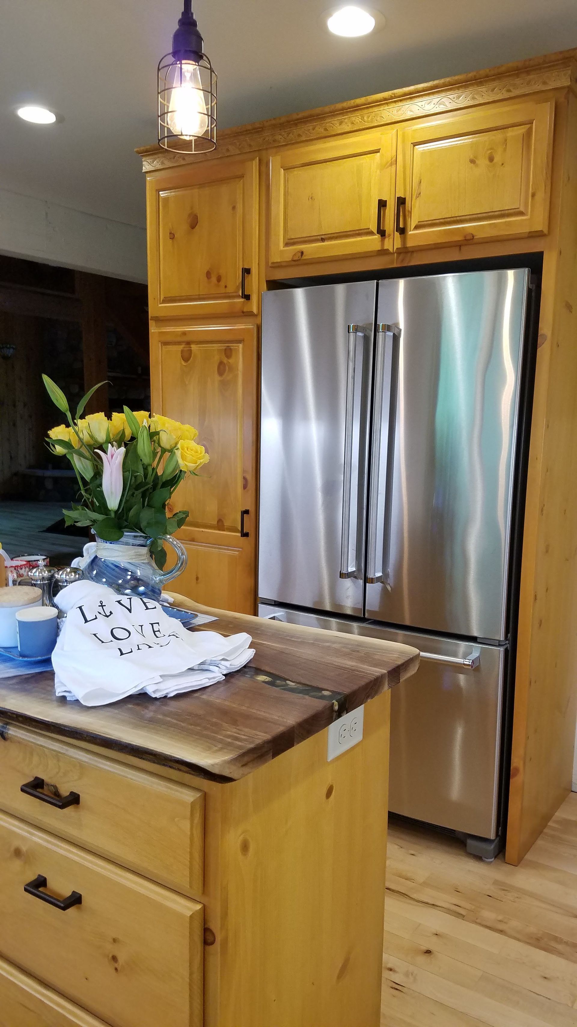 Kitchen with stainless steel refrigerator, light wood cabinets, and a countertop holding flowers and folded laundry