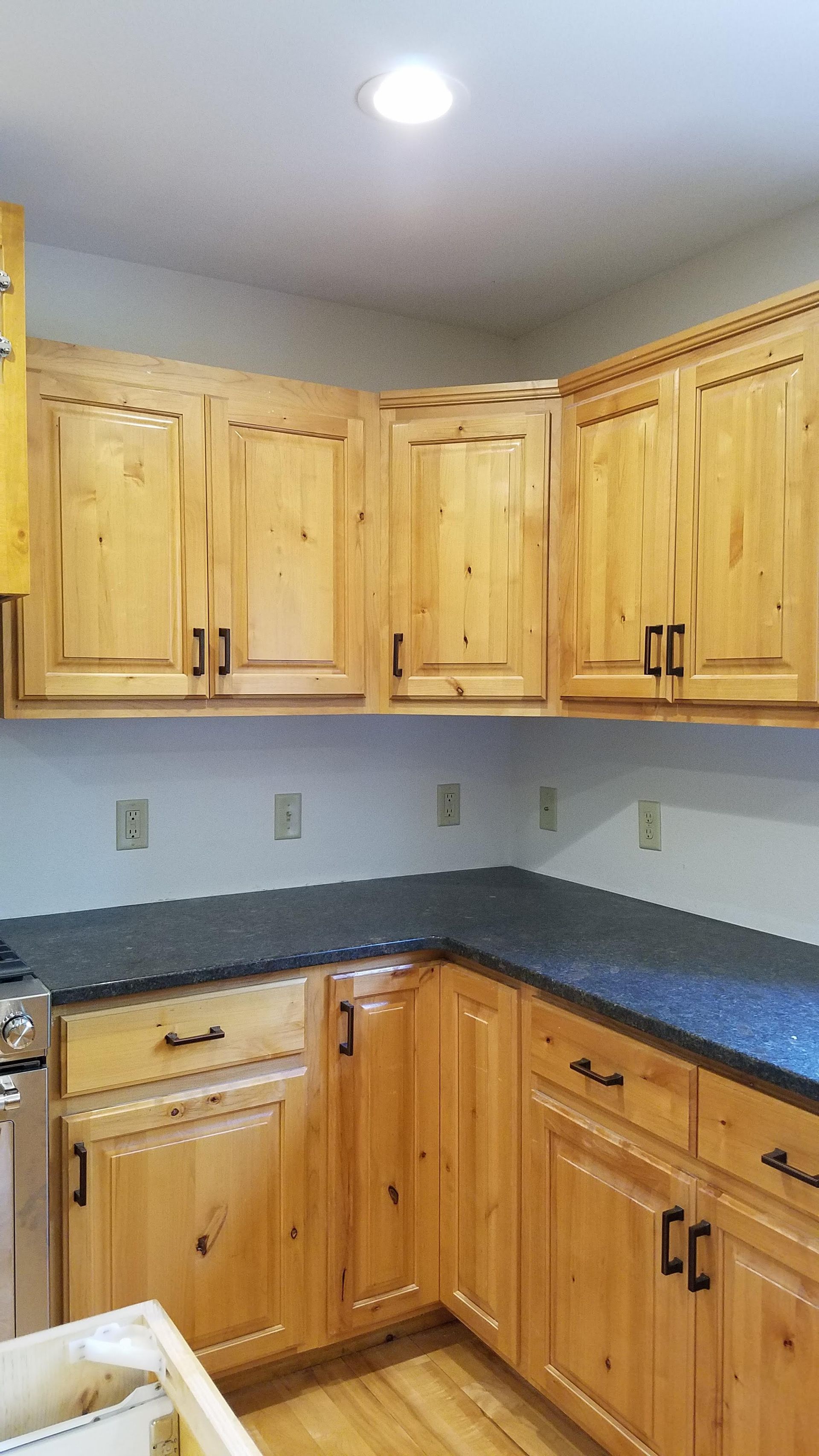Corner kitchen with light wood cabinets, dark countertop, and white walls under a ceiling light
