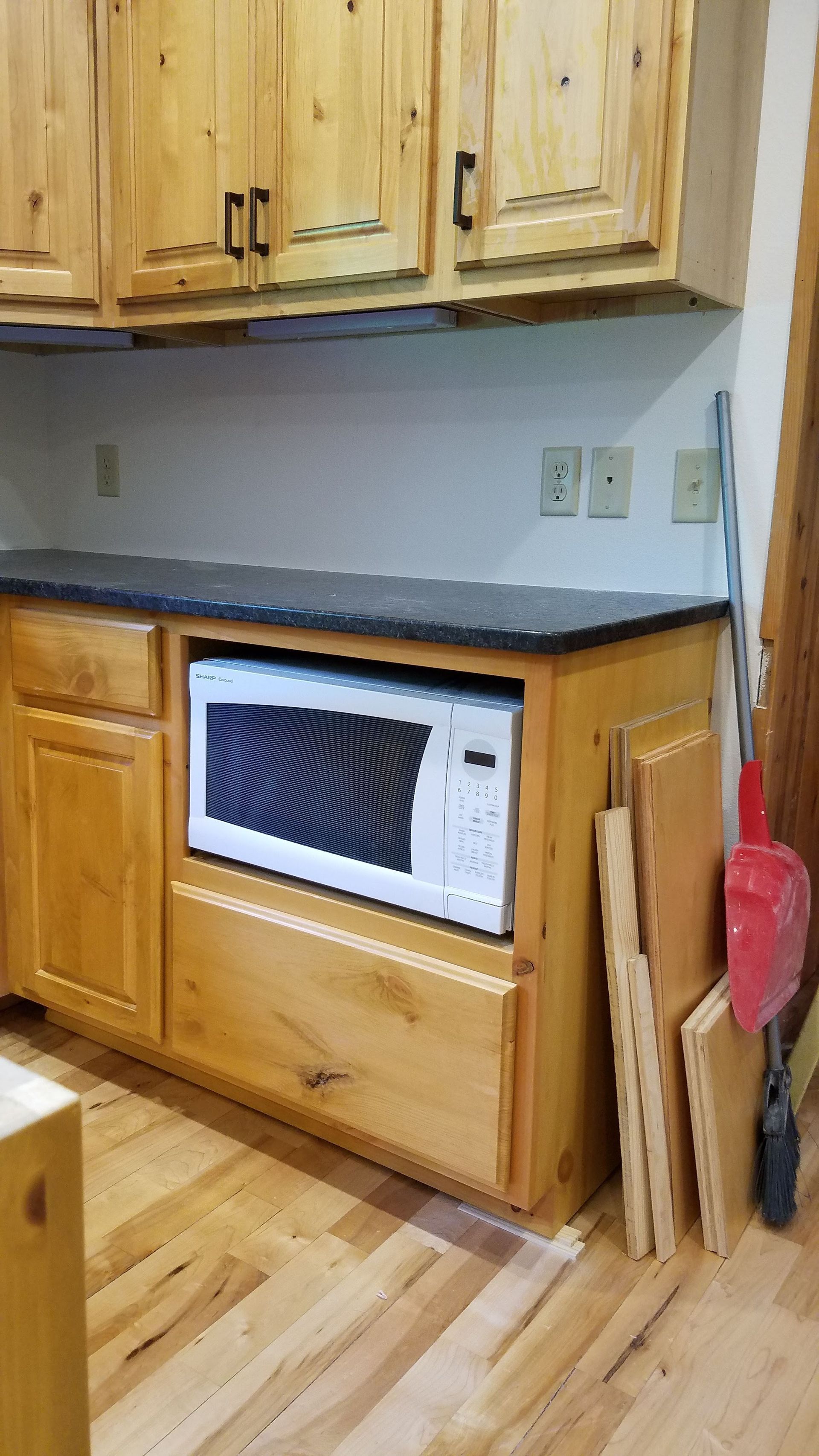 Kitchen cabinets with a built-in microwave under a dark countertop and wood floor, plus cleaning tools at right