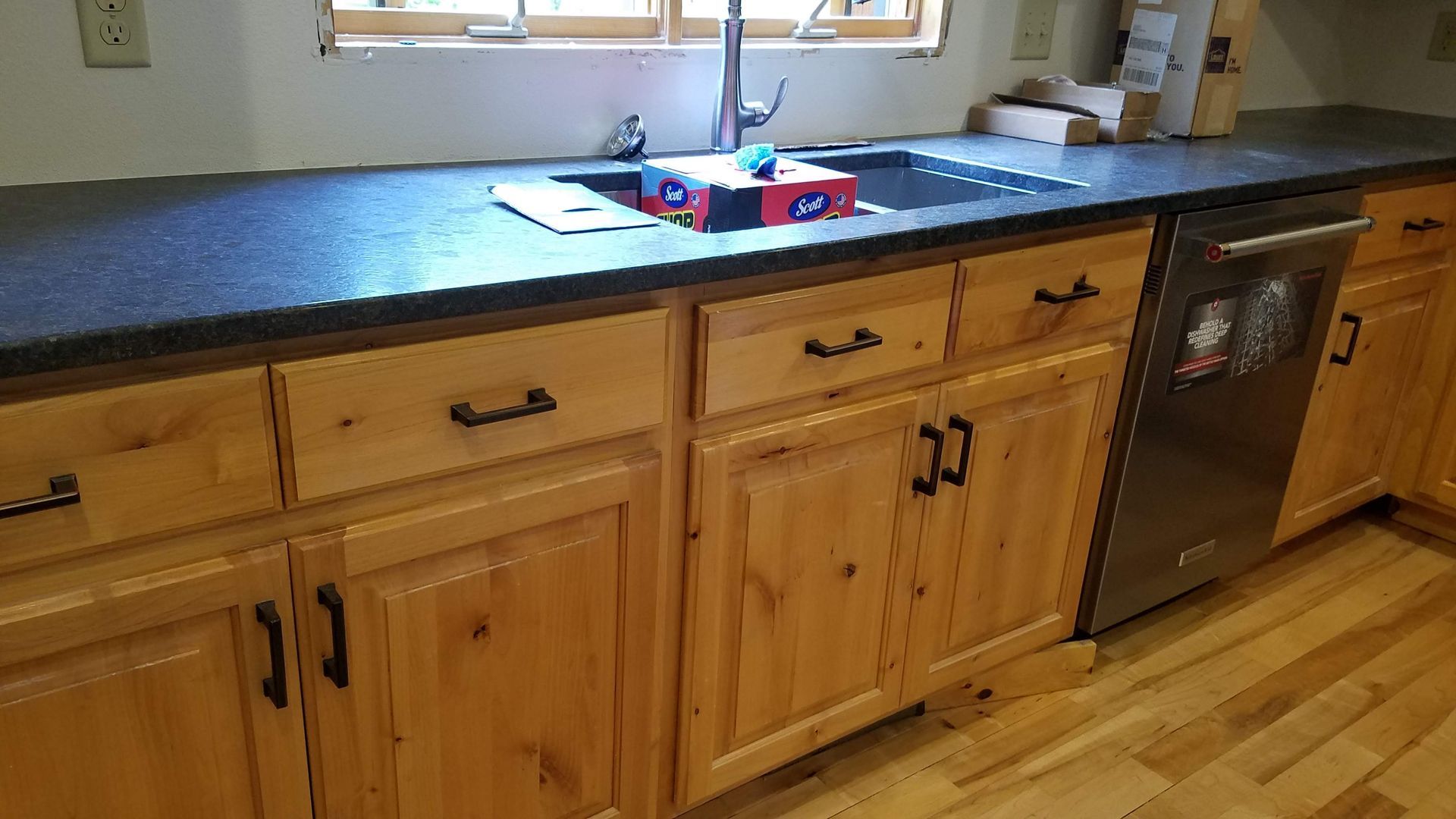 Wooden kitchen cabinets with a dark countertop, sink under a window, and a dishwasher on the right.