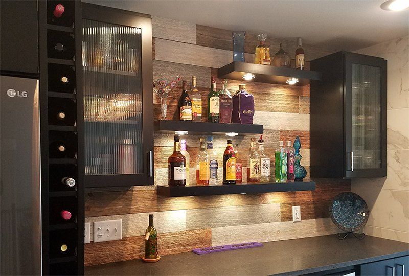 Home bar with shelves of liquor bottles, glass cabinets, and warm under-shelf lighting on a wooden wall.