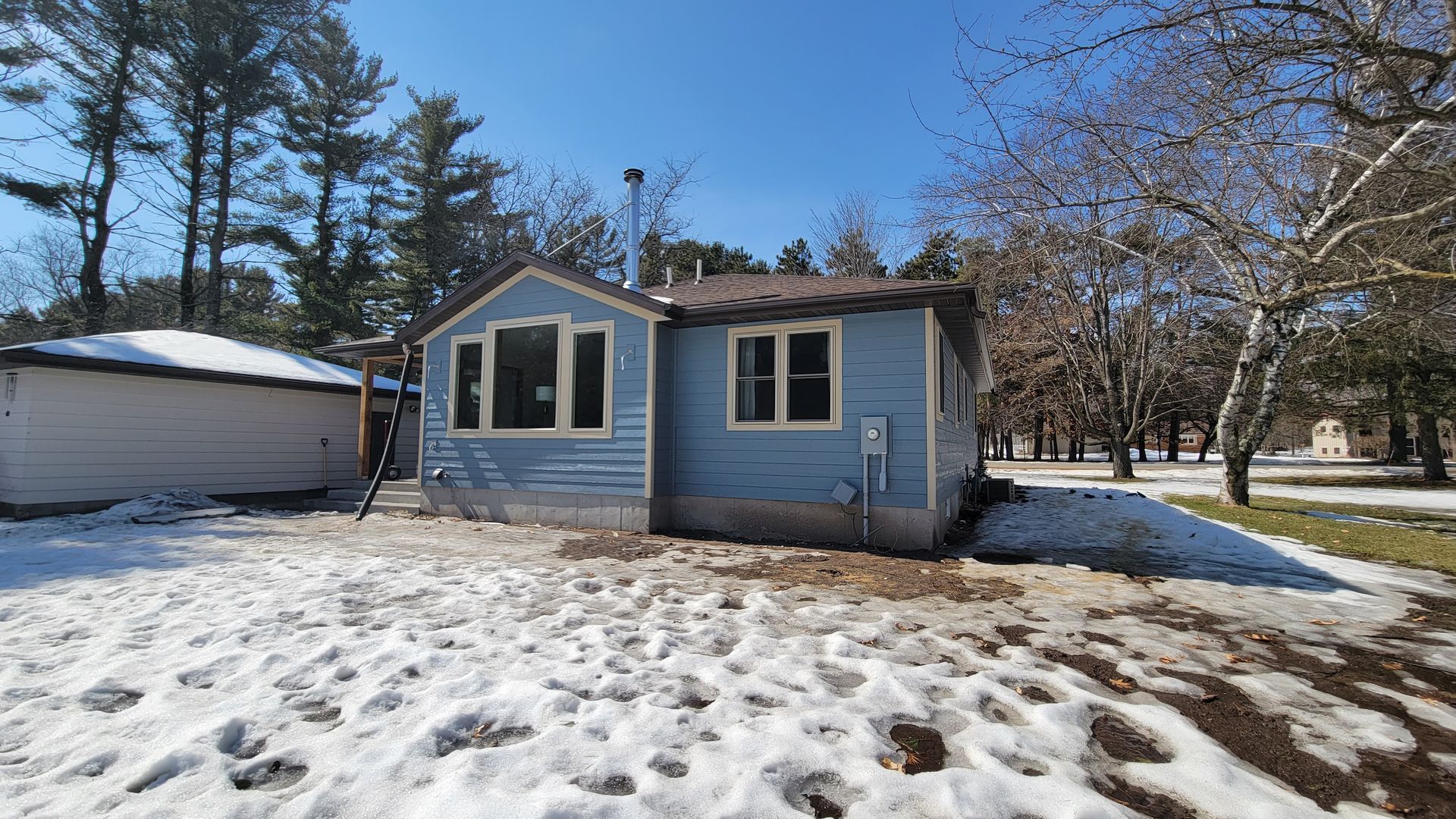 Small blue house with snow-covered ground and leafless trees under a bright sky.