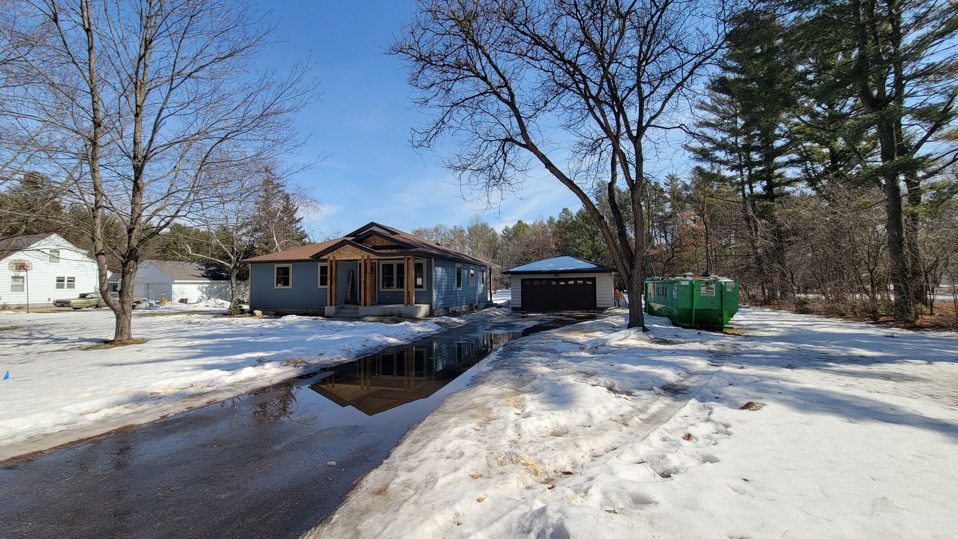 A snow-covered residential scene with a blue house, driveway, garage, and trees under a clear blue sky.