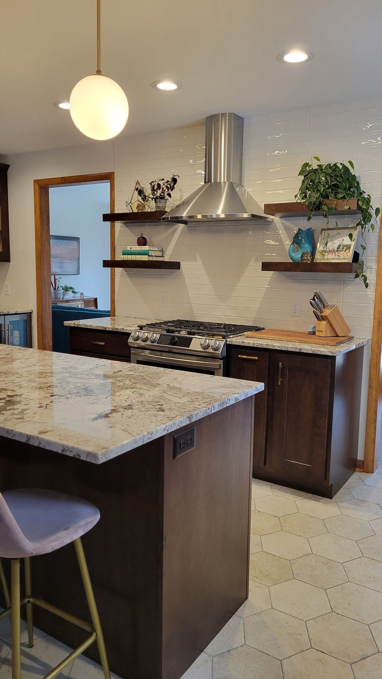 Kitchen with granite island, stove, stainless steel range hood, floating shelves, and globe light.