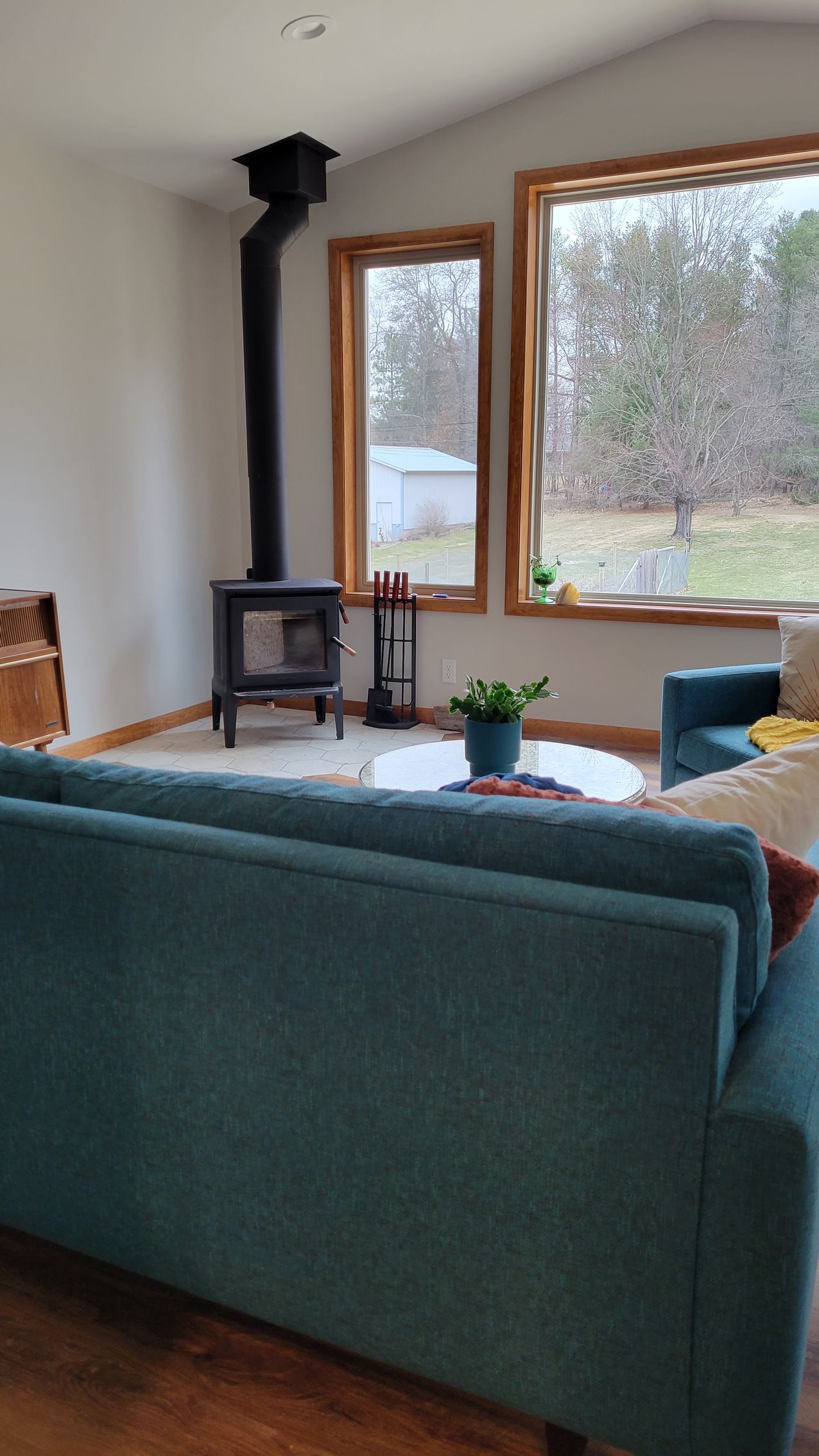 Living room with teal sofa, wood-burning stove, large windows with a view of trees.