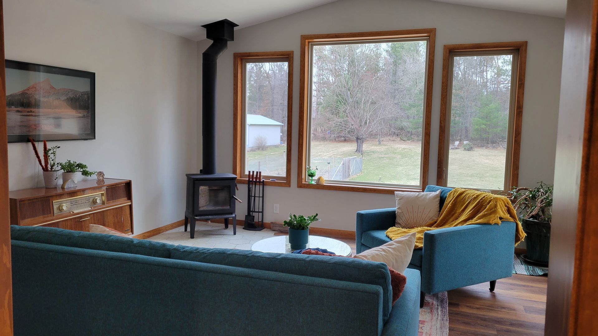 Living room with turquoise sofa and chair, fireplace, large windows overlooking yard, and art on the wall.