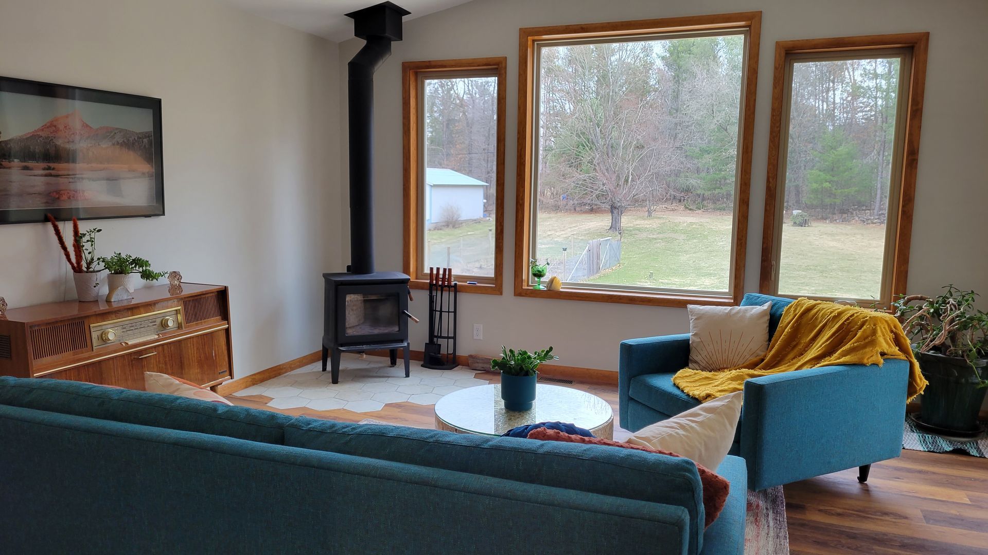 Living room with teal furniture, wood stove, round coffee table, and large windows overlooking a yard.