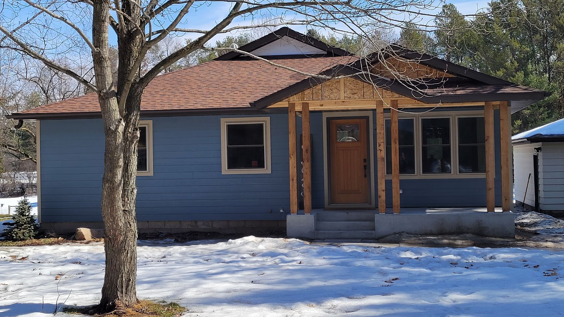 Blue house with brown roof, porch and door, snow on ground, tree in front.