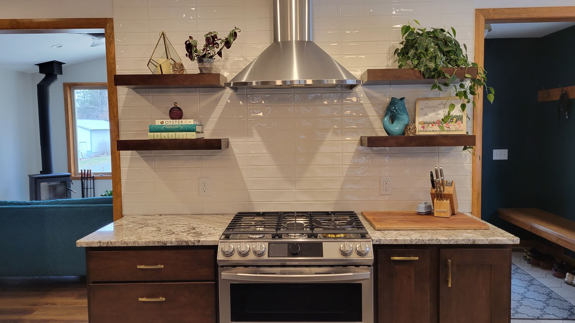 Kitchen with stainless steel range and hood, floating shelves with decor, brown cabinets, and white backsplash.