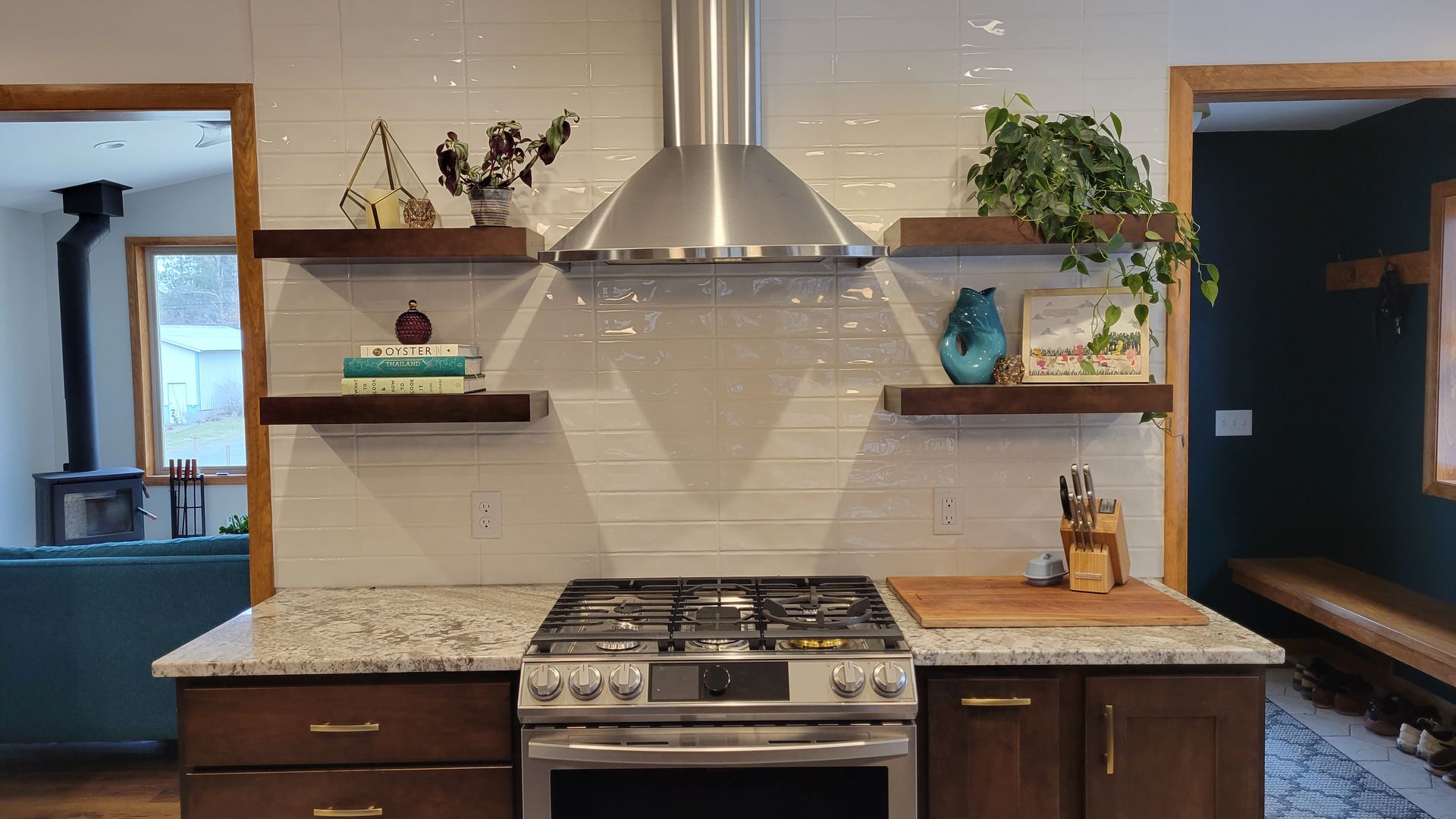 Kitchen with stainless steel range hood, wooden shelves, and dark cabinetry. White brick-like backsplash.