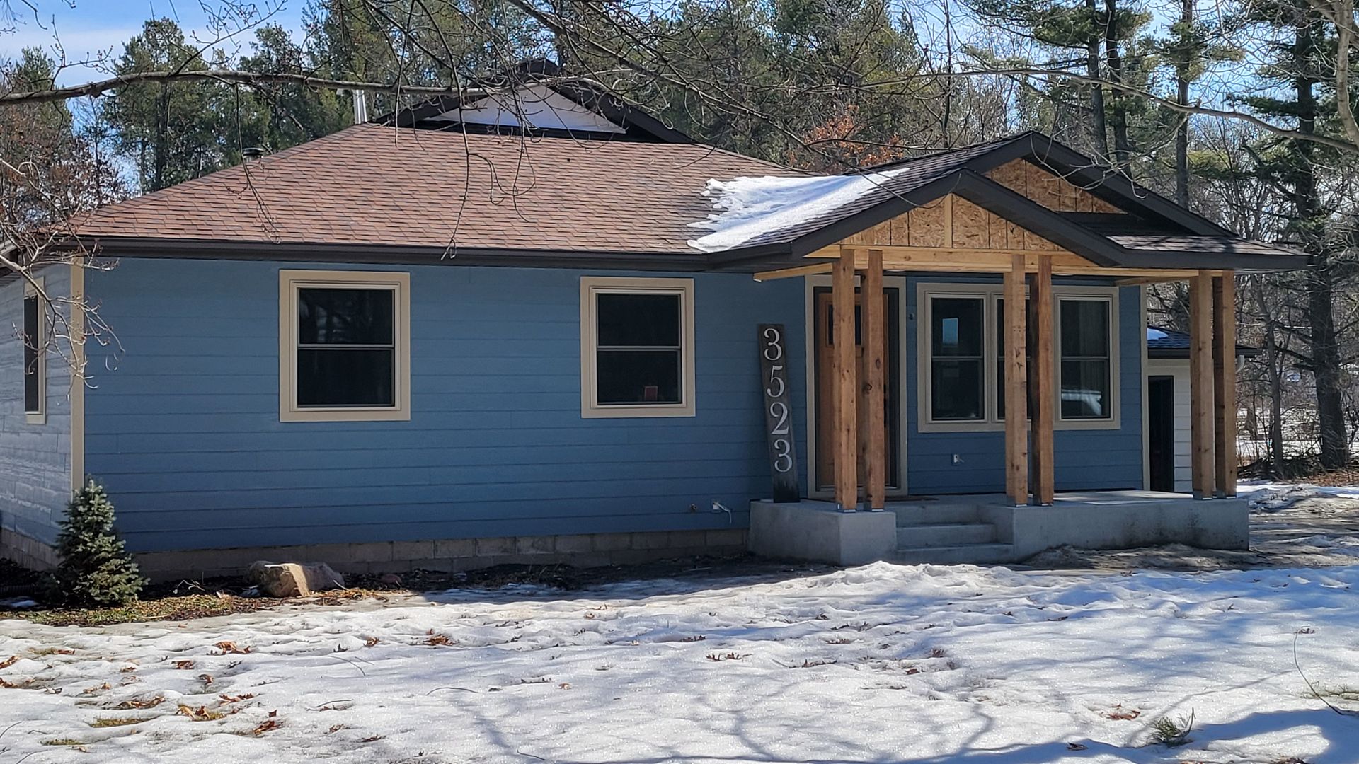 Blue house with brown roof and wooden porch, set in a snowy yard.