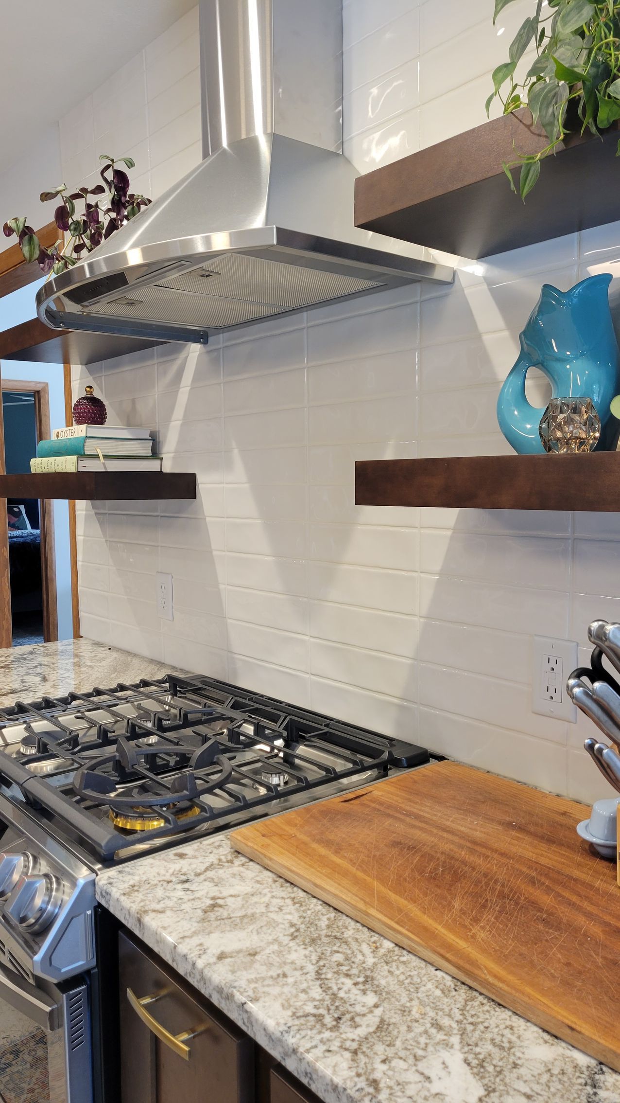 Kitchen with stainless steel range hood, floating shelves, white tiled backsplash, gas stovetop, and wooden cutting board.