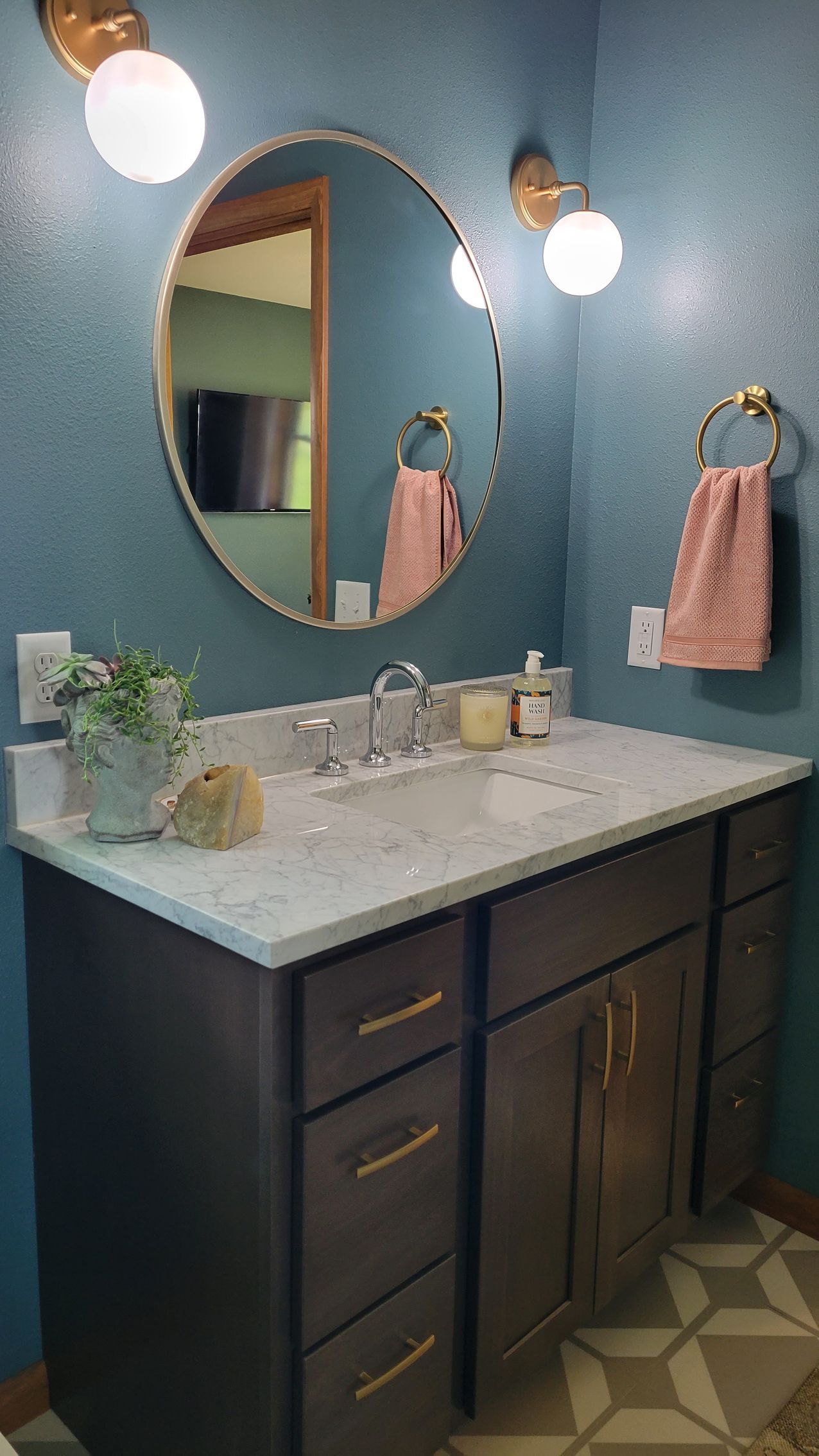 Bathroom with gray vanity, round mirror, blue walls, gold accents, pink towel.