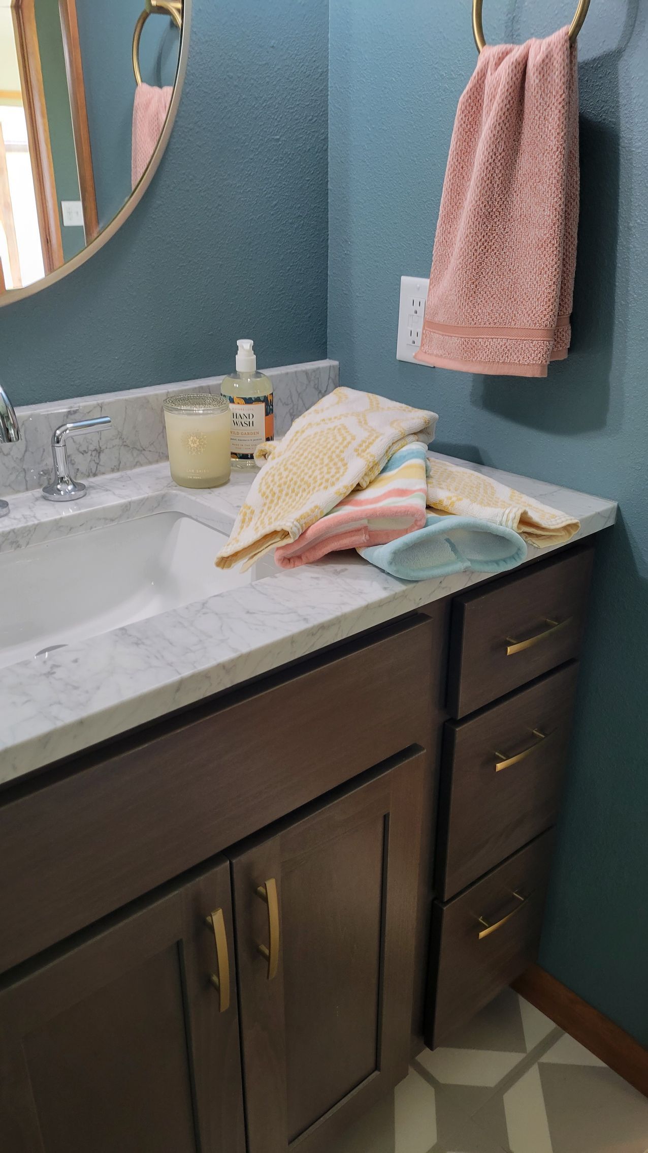 Bathroom vanity with towels. Gray cabinet, white countertop, teal wall, gold mirror. Pink and yellow towels.