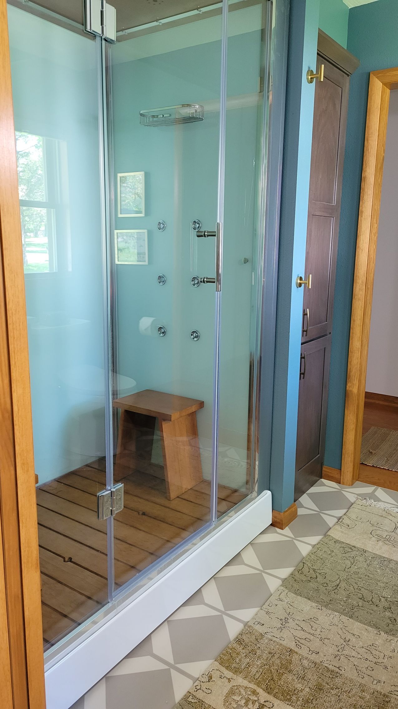 Shower with glass doors and wooden floor, featuring multiple shower heads and a small wooden stool.