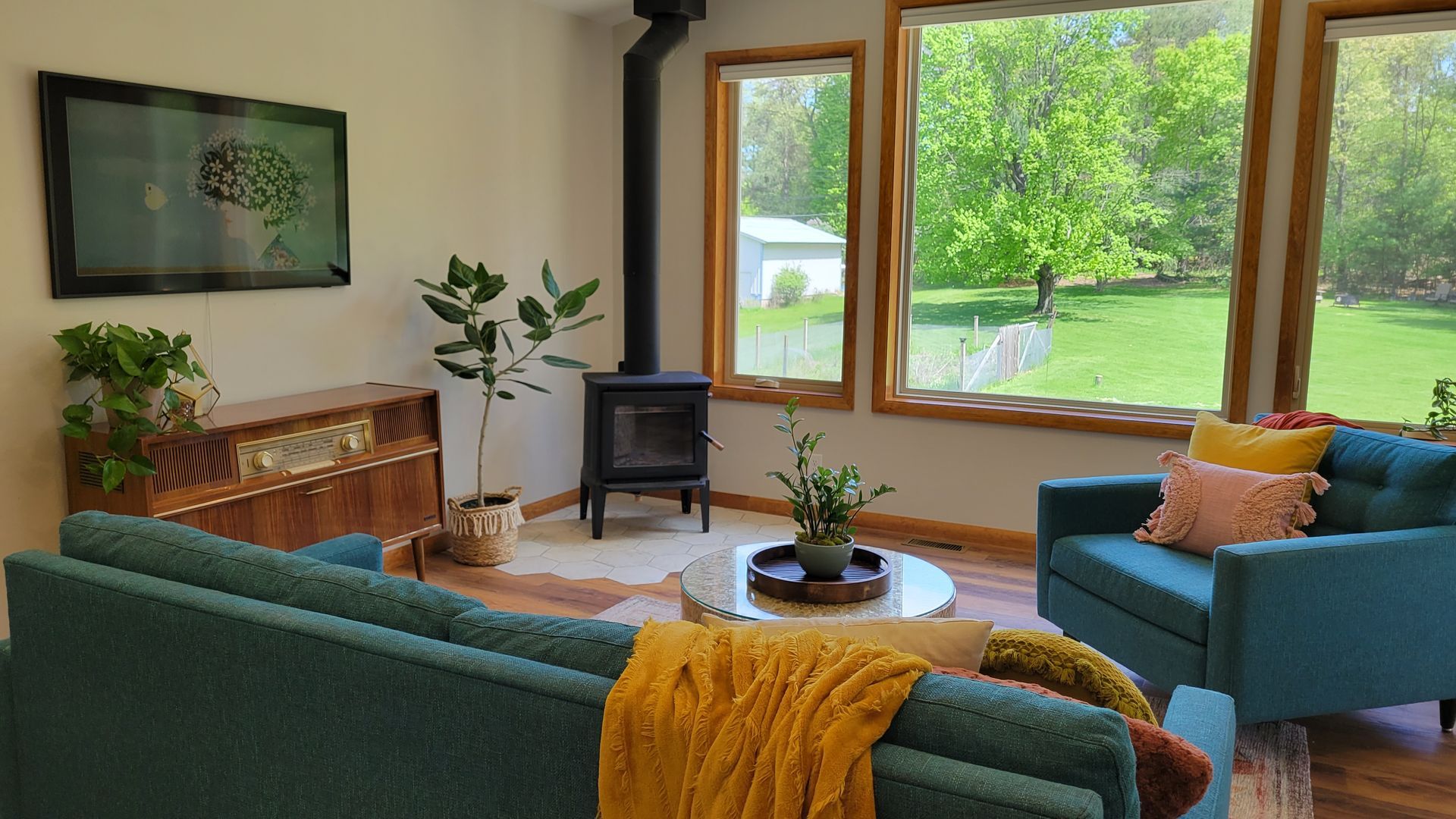 Living room with teal sofa and armchair, wood-burning stove, large windows, and outdoor view.
