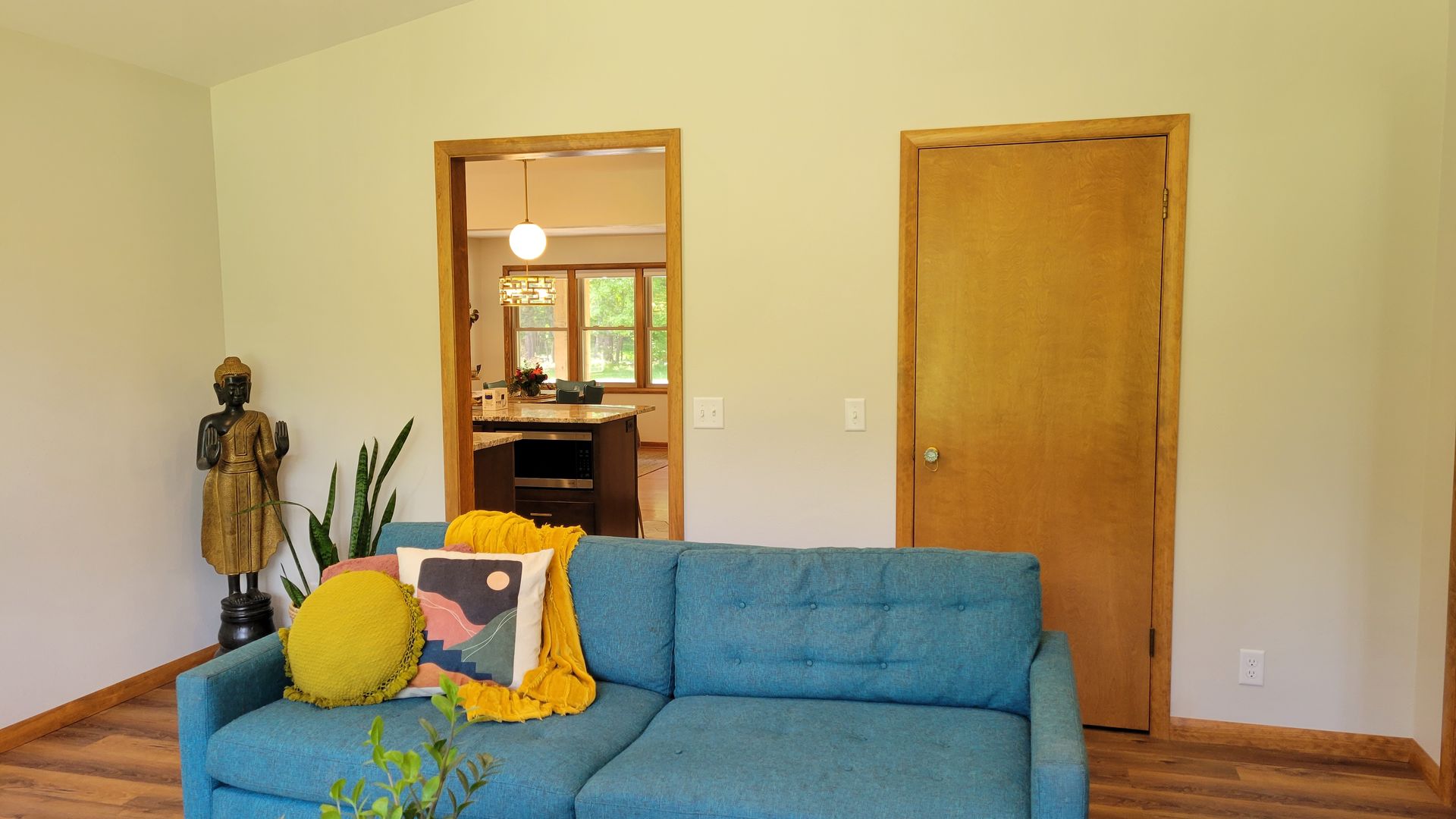 Teal couch in a living room with wooden floors. Doorway to kitchen, wooden door, statue and plants.