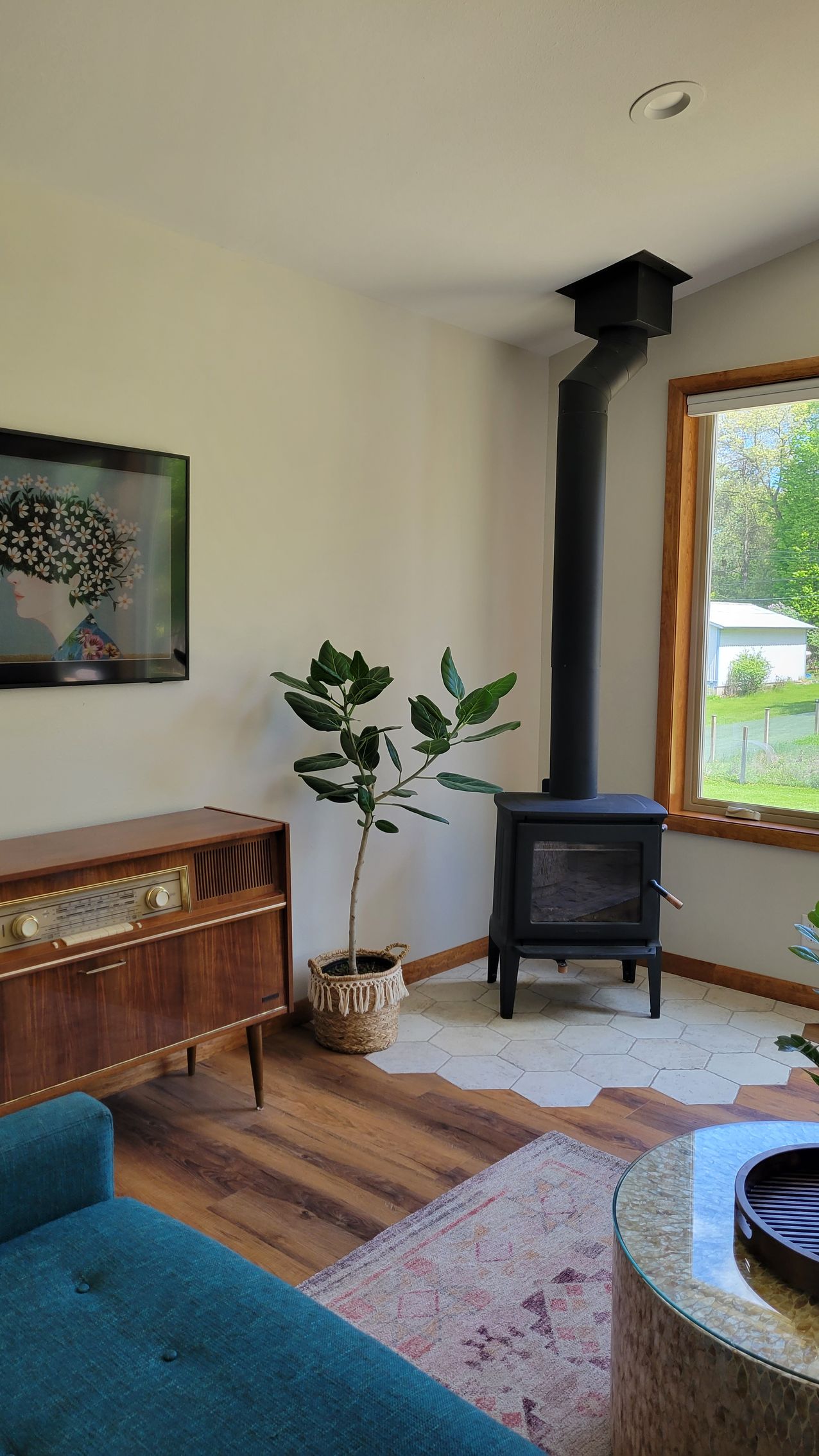 Living room with wood-burning stove, green plant, mid-century cabinet, teal sofa, and window.