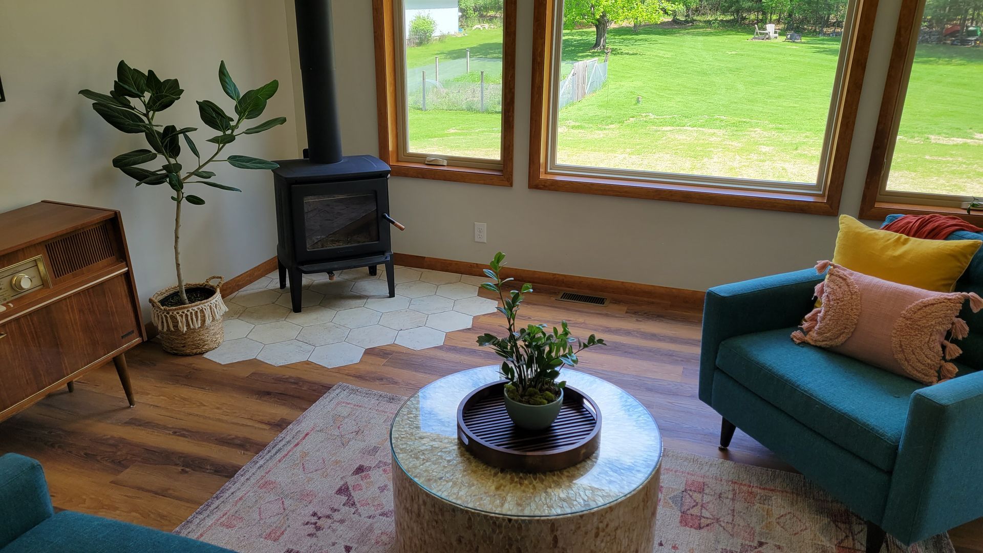 Living room with teal chairs, wood stove, tree stump coffee table, and large windows overlooking a green yard.