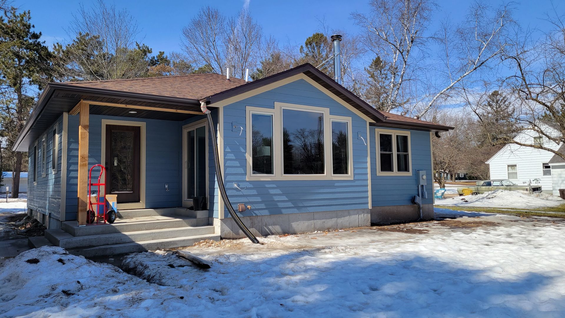 Blue house with brown roof and light trim, surrounded by snow and trees on a sunny day.