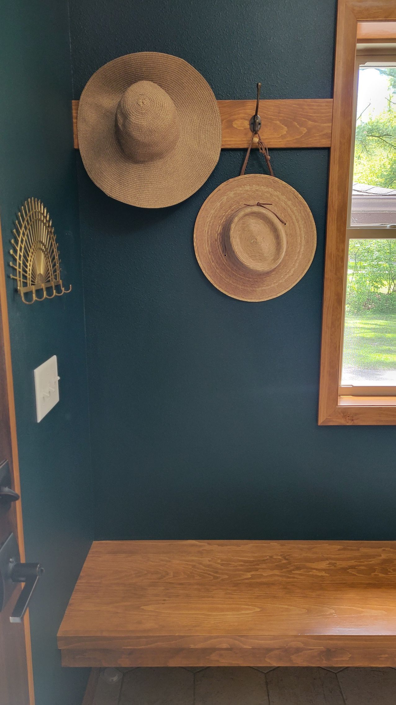 Two straw hats hang on a wooden rack above a wooden bench, set against a teal wall next to a window.
