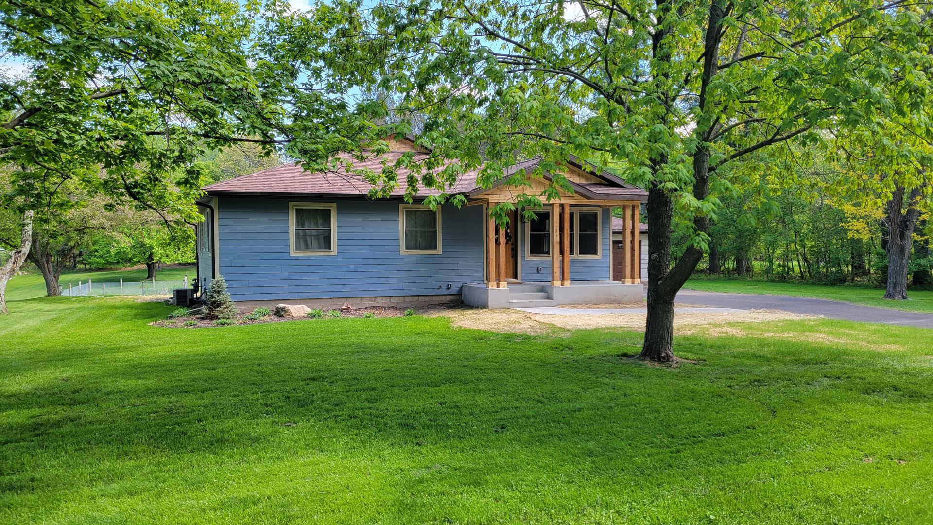 Blue house with brown roof, green grass, and trees. Front porch has tan columns. Sunny day.