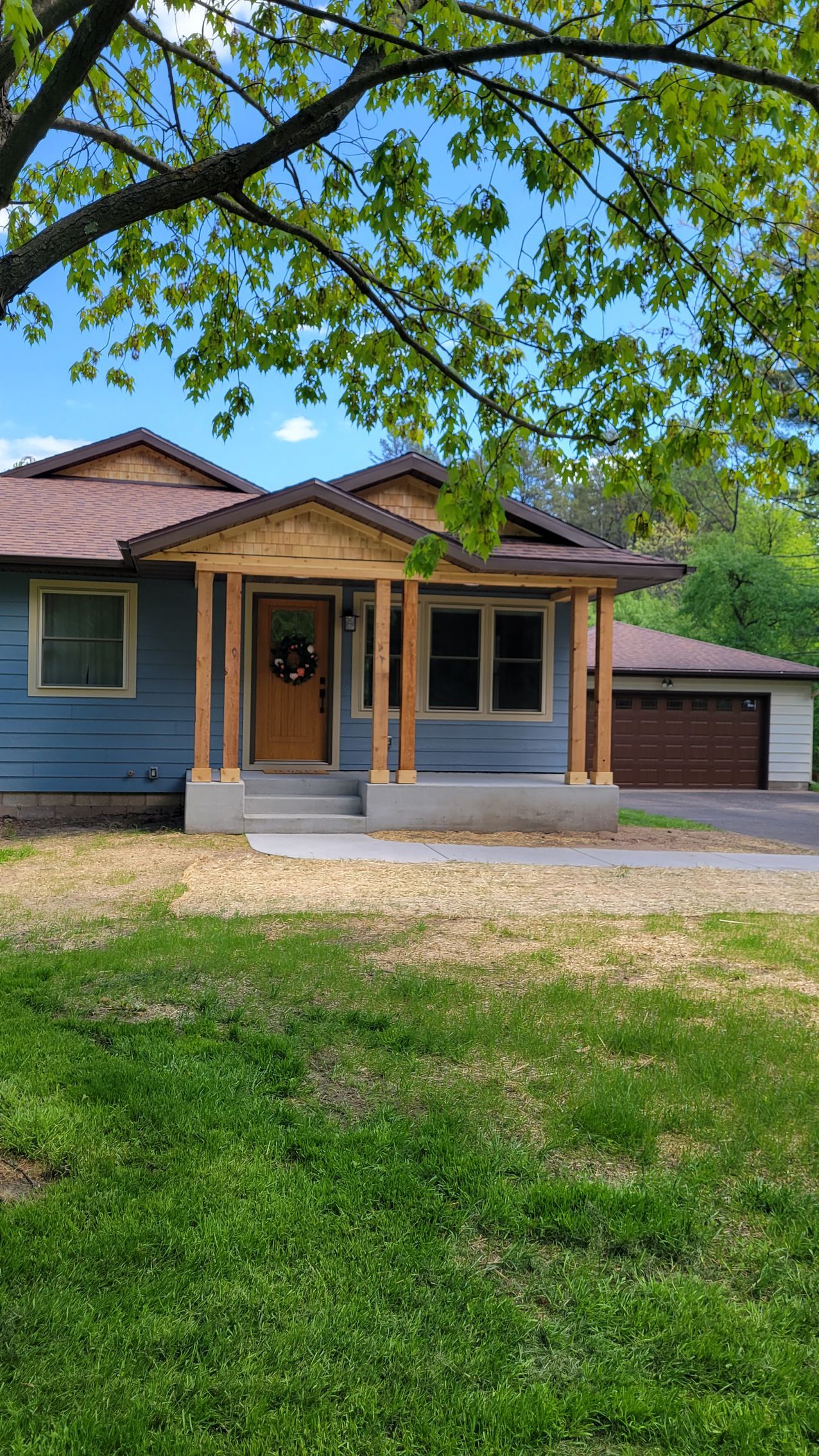 Blue house with wooden porch and garage; green lawn and tree overhead.