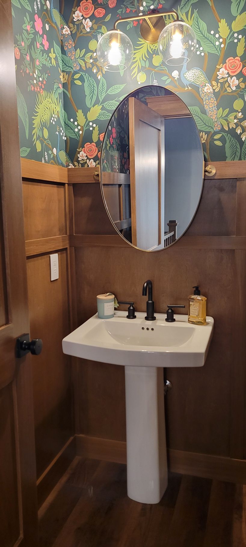 A bathroom with floral wallpaper, an oval mirror, and a white pedestal sink.