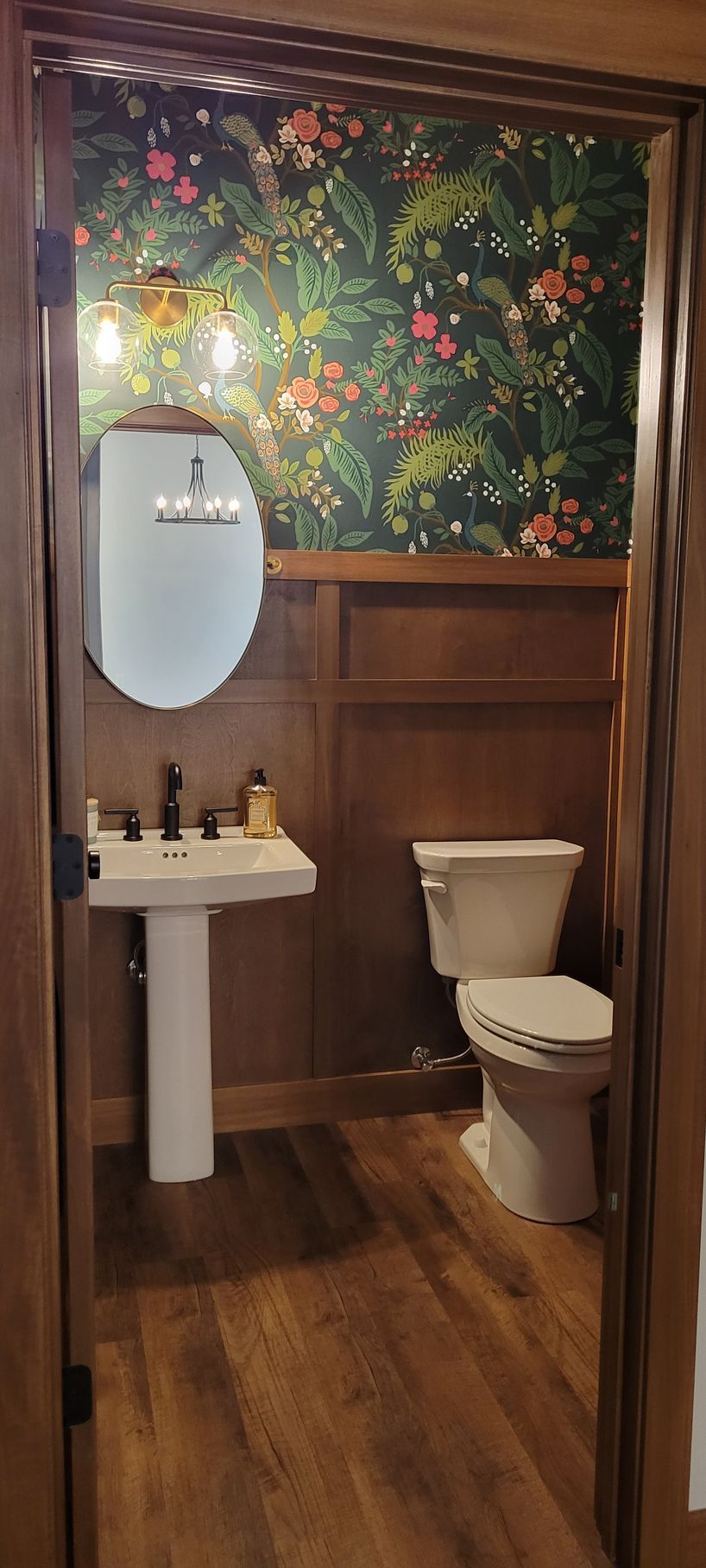 A small bathroom with floral wallpaper, brown wood paneling, a pedestal sink, and a toilet.