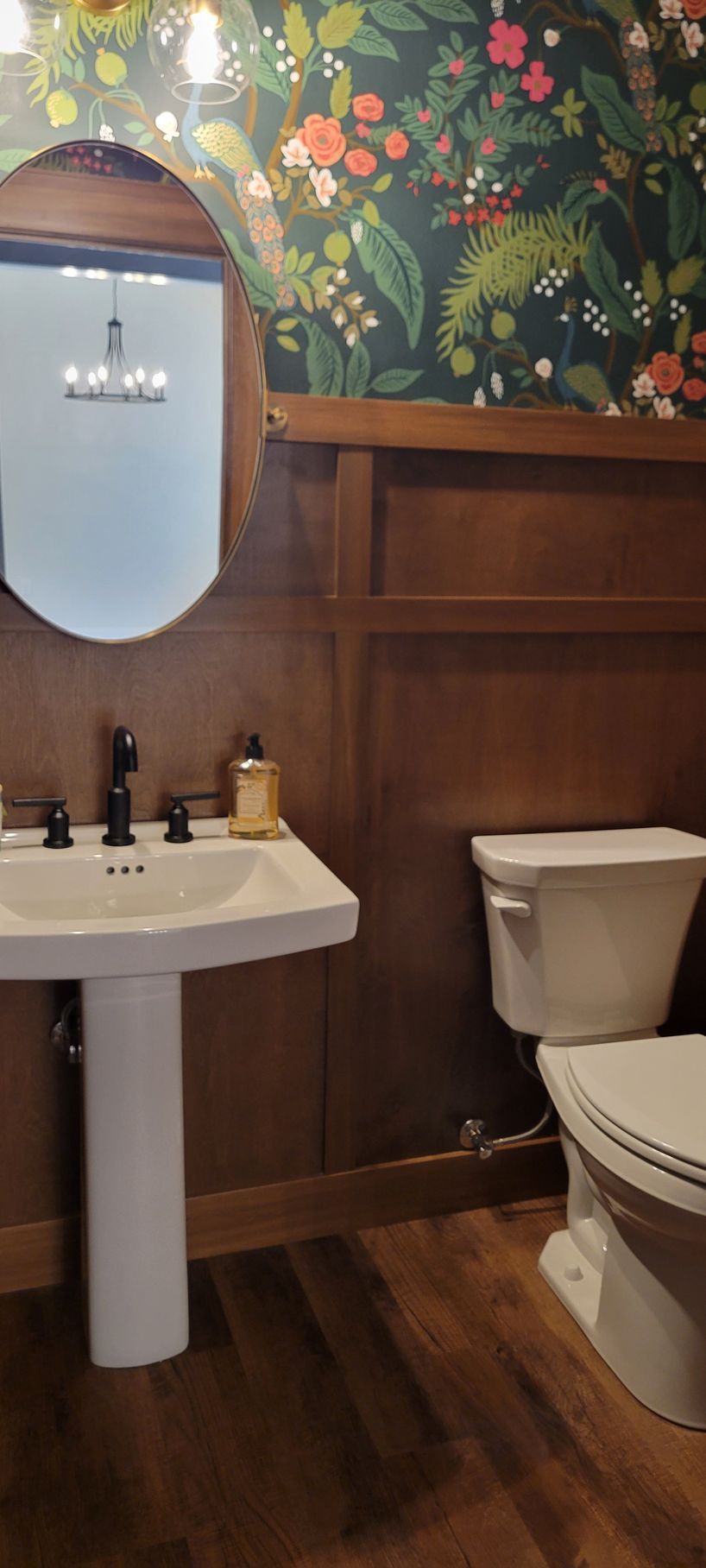 Bathroom with pedestal sink, toilet, dark wood paneling, and floral wallpaper.